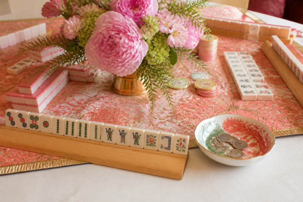 Close-up of an elegantly set mahjong game table with bamboo racks, colorful tiles arranged by suit, a mahjong money dish with red and green dragon design holding coins, a festive coral and gold table runner, and a fresh floral centerpiece with pink peonies and green foliage, shot under warm afternoon lighting