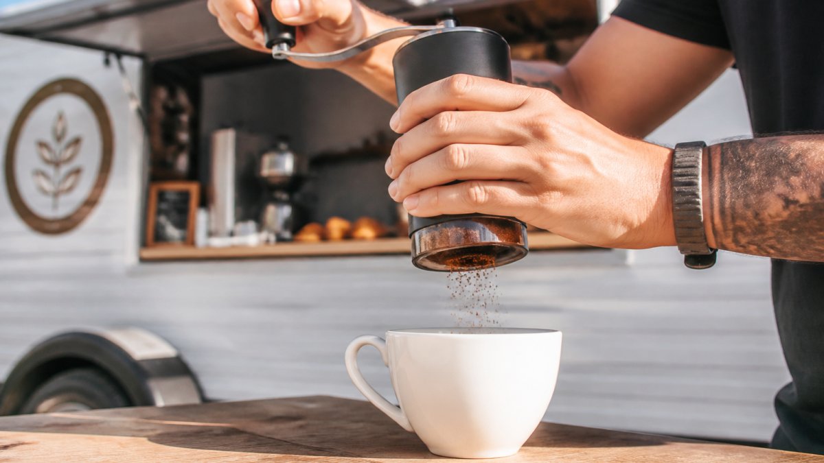 Hands holding a manual coffee grinder over a white ceramic cup, showing the grinding motion and the adjustment ring detail with natural morning light