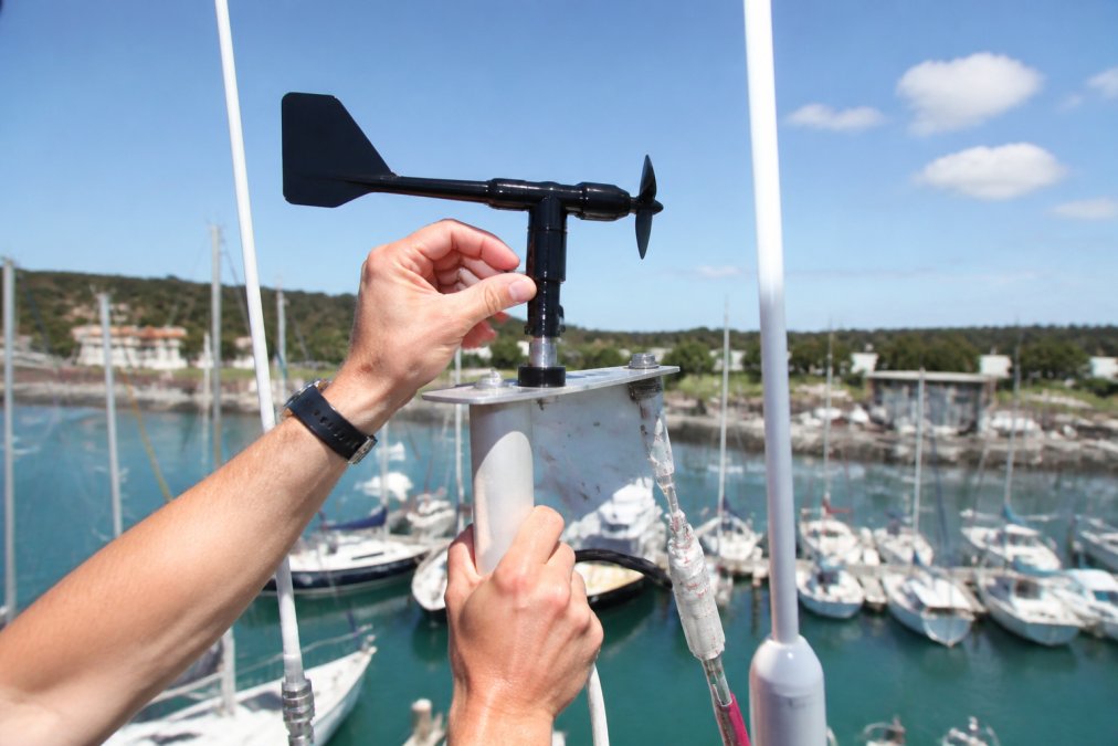 A sailor's hands attaching a Raymarine wind transducer to a masthead bracket high above a marina, blue sky and rigging lines visible, bright midday light
