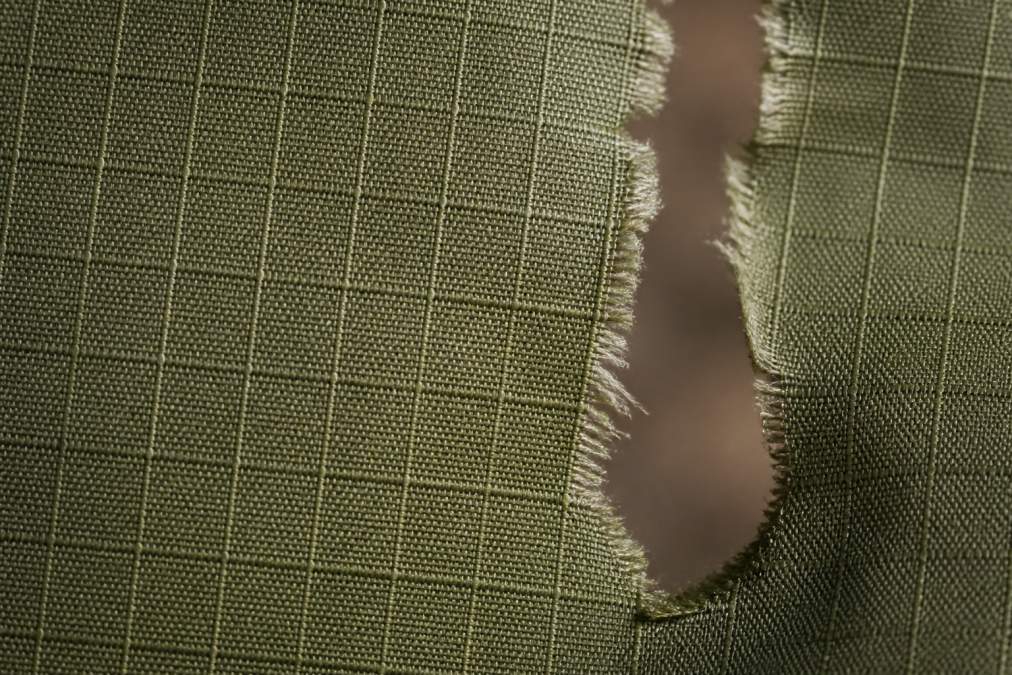 Close-up of ripstop weave fabric showing the reinforced grid pattern with visible fibers under natural light, with a torn edge demonstrating how the grid stops tear propagation