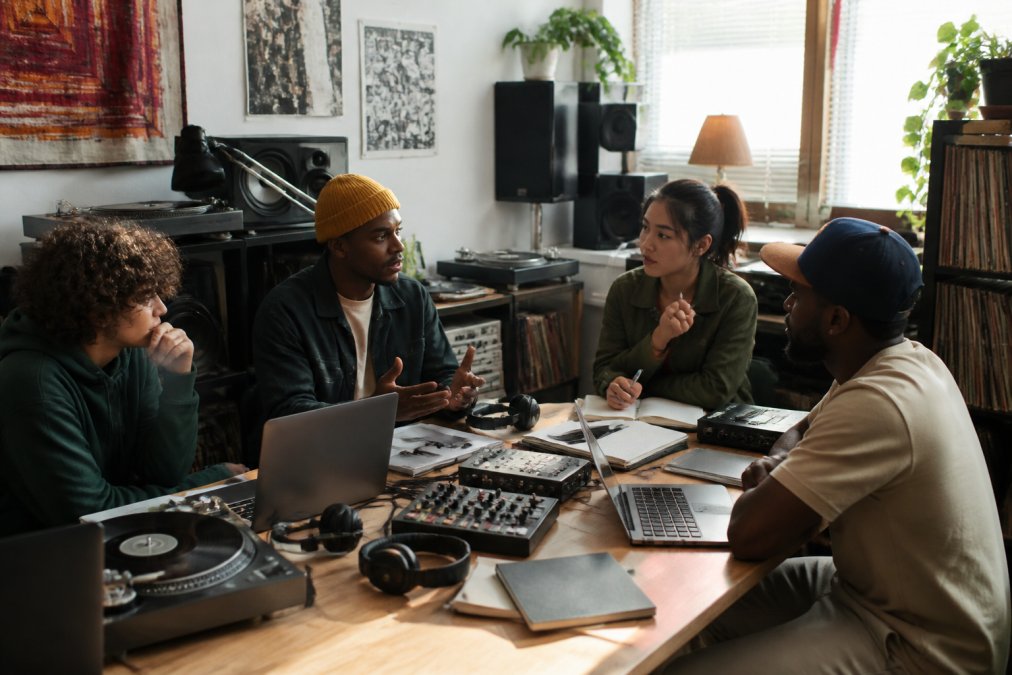 Independent music entrepreneur collaborating with team members and artists around a desk with laptops, headphones, and mixing equipment in a modern home studio environment with natural window lighting