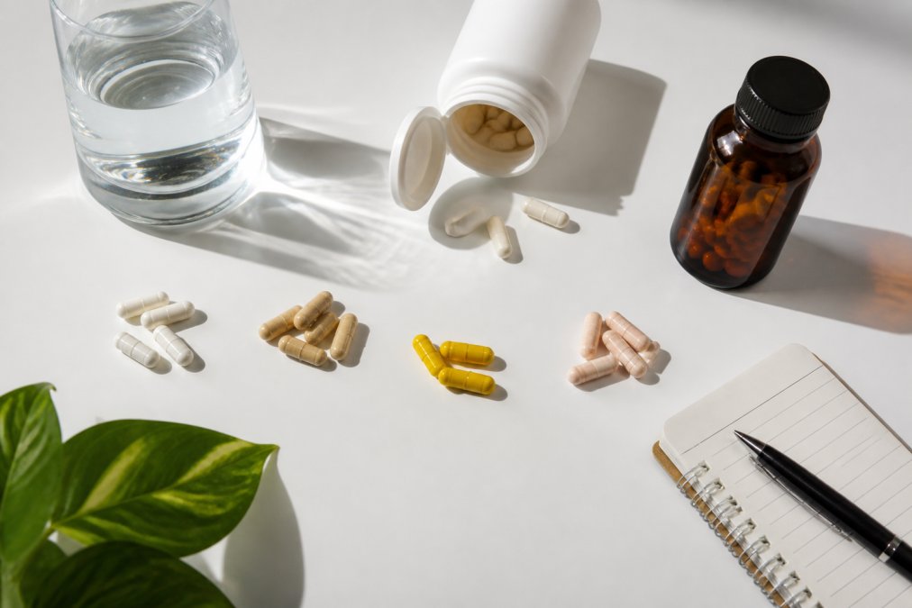 Bright flat-lay photo of supplement bottles and capsules arranged on a clean white table with a glass of water and a small notepad, natural daylight
