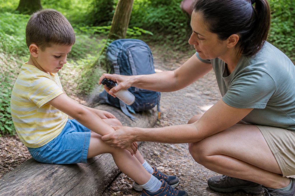 Parent carefully applying natural tick repellent spray to child's exposed arms, legs, and socks before outdoor hiking, showing proper technique with even coverage in natural daylight