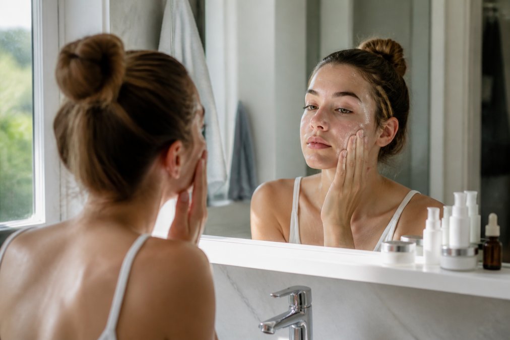 A teenager applying lightweight moisturizer to clean, damp face in a bathroom mirror, with skincare products visible on the counter in natural morning light