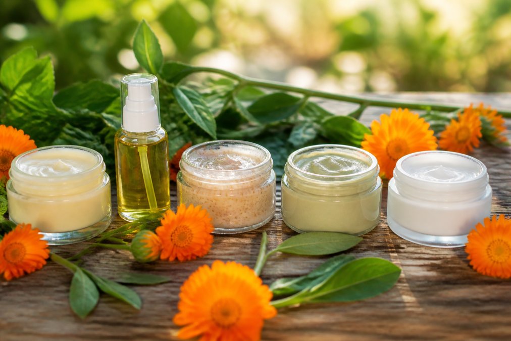 A flat lay of five different organic cleansers in glass and recyclable jars on a rustic wooden table, surrounded by fresh calendula flowers and green leaves, soft morning light casting gentle shadows