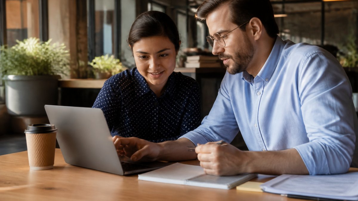 A startup founder and office manager reviewing payroll options on a laptop in a bright coworking space, notebook and coffee on the desk, natural window light
