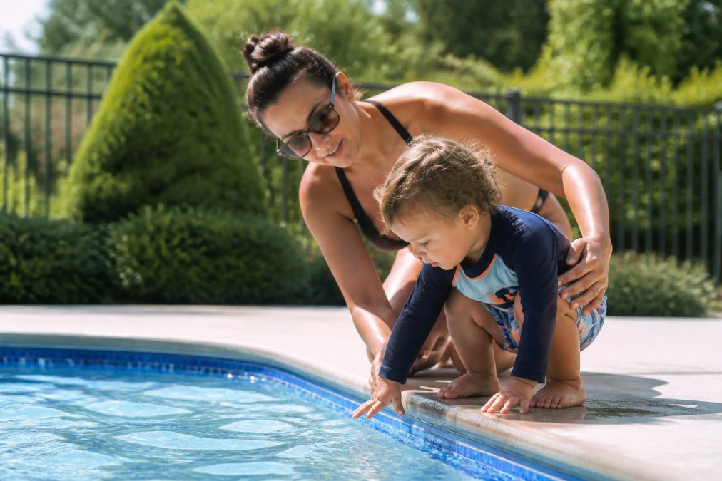 Parent and young toddler at the pool's edge with parent's hand gently guiding child, showing attentive supervision in bright afternoon sunlight