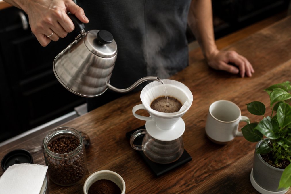 Hands pouring hot water from a gooseneck kettle into a ceramic pour over coffee dripper on a wooden counter