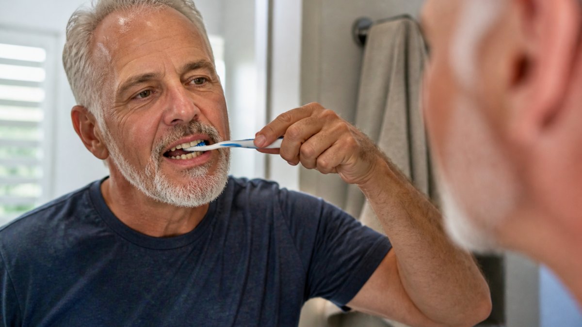 Close-up of hands demonstrating proper soft-bristled toothbrush technique at 45-degree angle to gum line around implant-supported crown in clinical setting with natural lighting