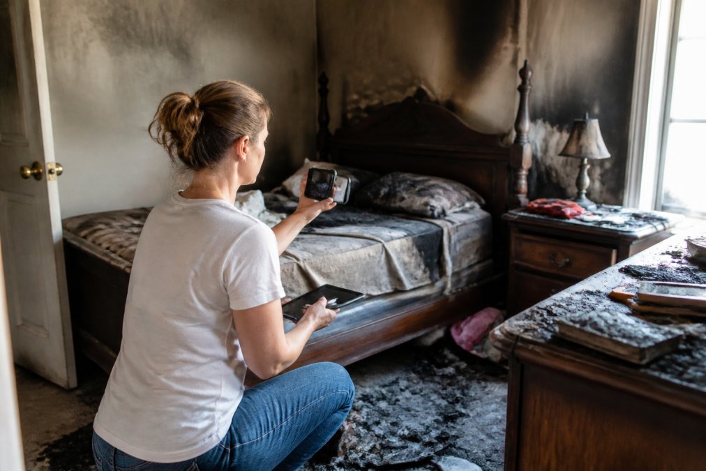 Homeowner photographing household items and taking detailed notes on tablet while documenting personal property in bedroom, with damaged furniture and belongings visible in background