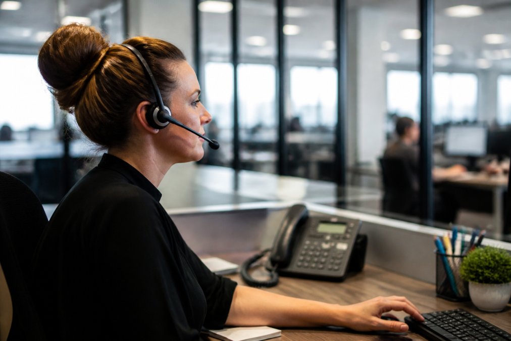 Dispatcher at desk with multiple monitors displaying real-time dispatch software, headset on, actively assigning emergency service jobs to field technicians across a service map with color-coded priority indicators and technician location pins
