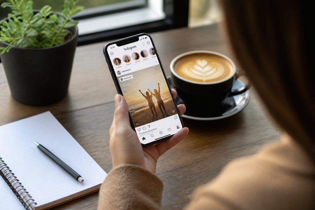 A close-up of a smartphone screen showing an Instagram feed with a reposted story, held by a person sitting at a wooden desk with a notebook and coffee nearby