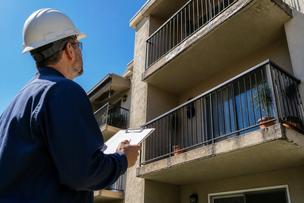 A property manager in a hard hat examining a multi-story apartment building balcony with a clipboard, inspecting exterior railings and structural elements on a sunny day