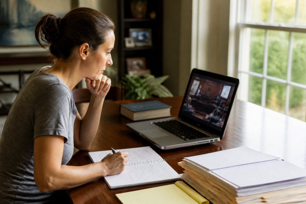 Person at home office on laptop taking online course with course materials and handwritten notes visible on desk, representing flexible remote learning in residential setting with natural window lighting