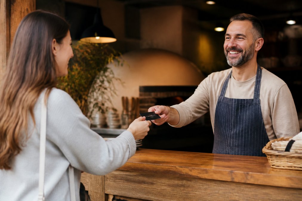 A smiling restaurant owner handing a membership card to a happy customer across a warm wooden counter in a cozy independent restaurant, soft overhead lighting illuminating their exchange