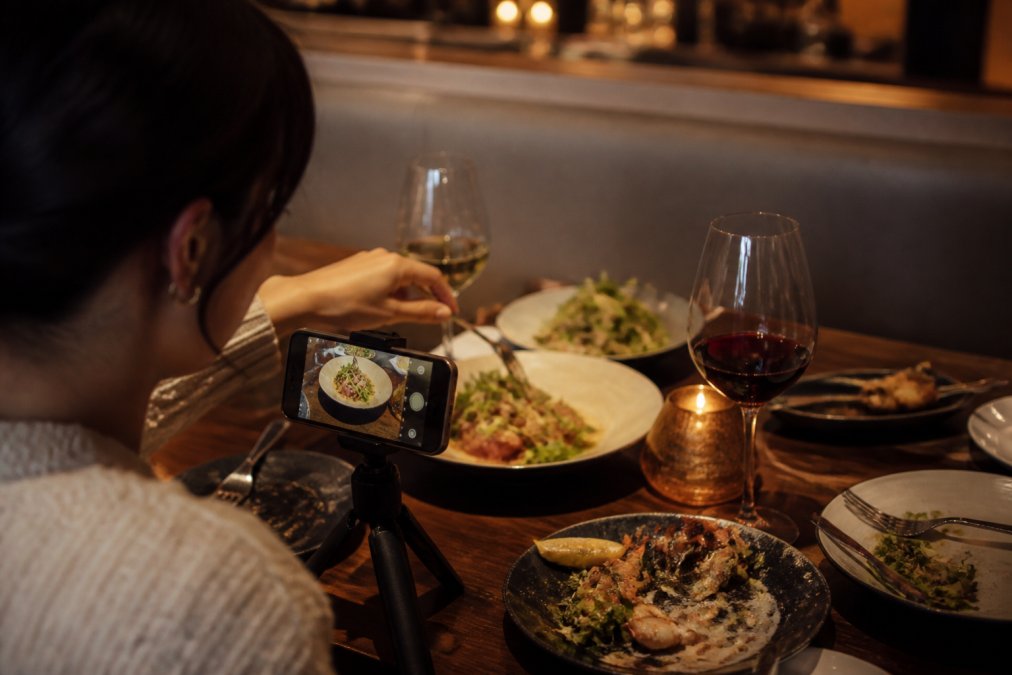 A food blogger photographing a beautifully plated dish at a restaurant table, smartphone mounted on a small tripod, warm amber ambient lighting in the background, half-eaten dishes and wine glasses visible nearby