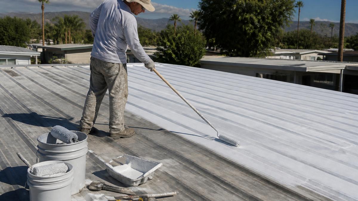 A worker on a metal mobile home roof applying silver reflective coating with a roller, sunny day, clean prepared surface visible, safety gear worn