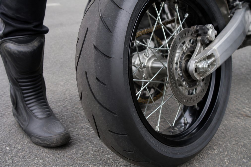 Close-up of a supermoto tire mounted on a 17-inch wheel showing tread pattern and sidewall markings in natural lighting, with a rider's boot nearby for scale