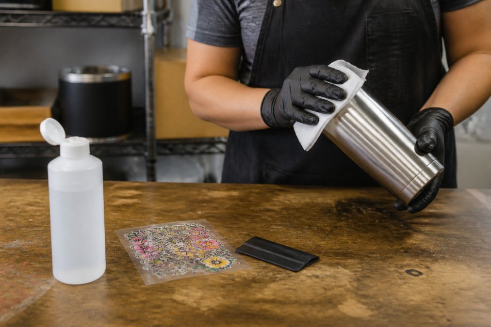 Cleaning a stainless steel tumbler with alcohol before applying UV DTF transfers on a workbench.