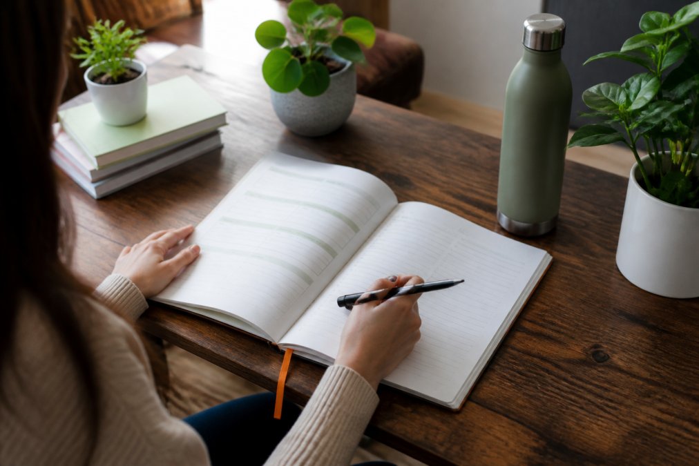 Person's hands holding open workbook with pen, sitting at desk with water bottle and plants visible, representing intentional living and habit tracking