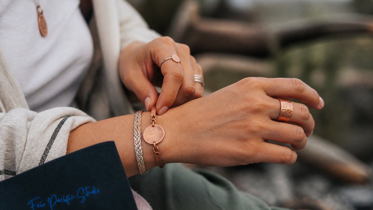 Close-up of hands wearing minimalist handcrafted jewelry with natural textures, showcasing sterling silver and reclaimed copper pieces against a soft, natural outdoor setting