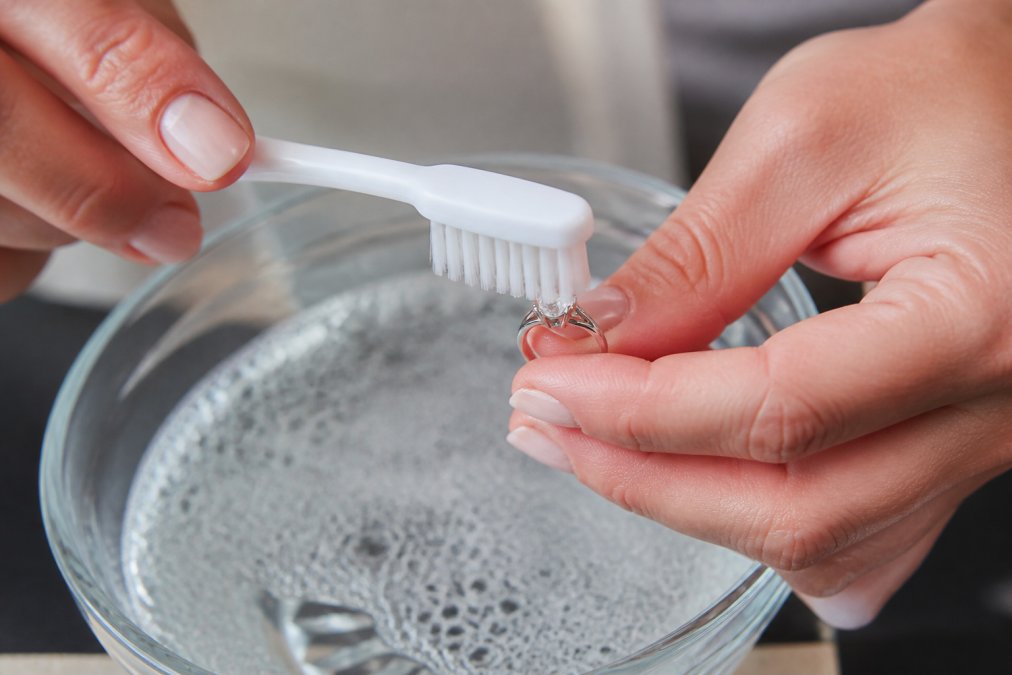 Close-up of hands gently cleaning an antique diamond ring with a soft-bristled toothbrush over a bowl of soapy water, showing delicate brushing motion around the prong settings in natural light