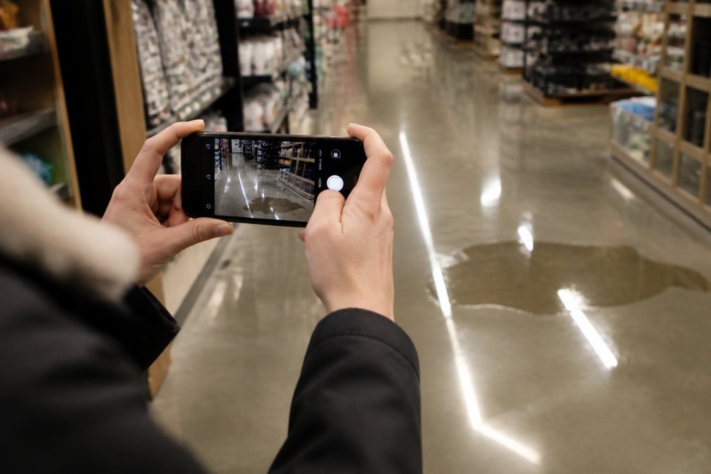 Close-up of a smartphone camera capturing a wet floor hazard in a retail store with water pooling visibly on the floor surface, hazard clearly in frame under bright overhead lighting