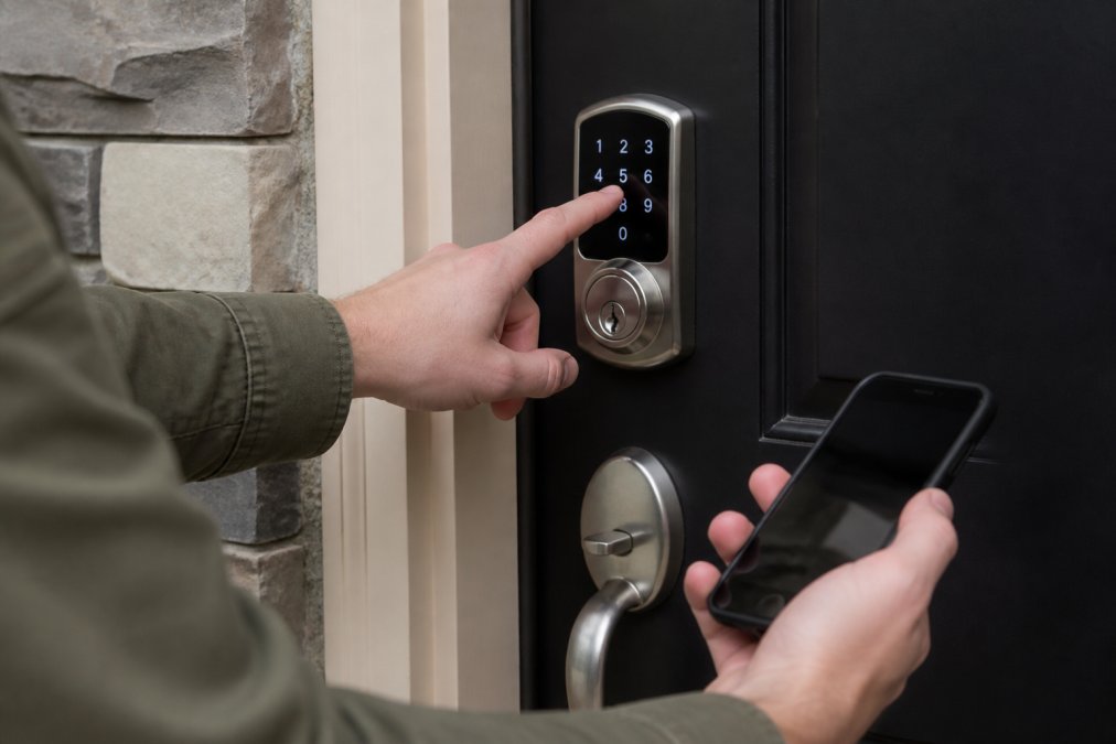 A homeowner's hand entering a code on a sleek, modern keypad deadbolt mounted on a front door, with a smartphone visible nearby showing the companion app