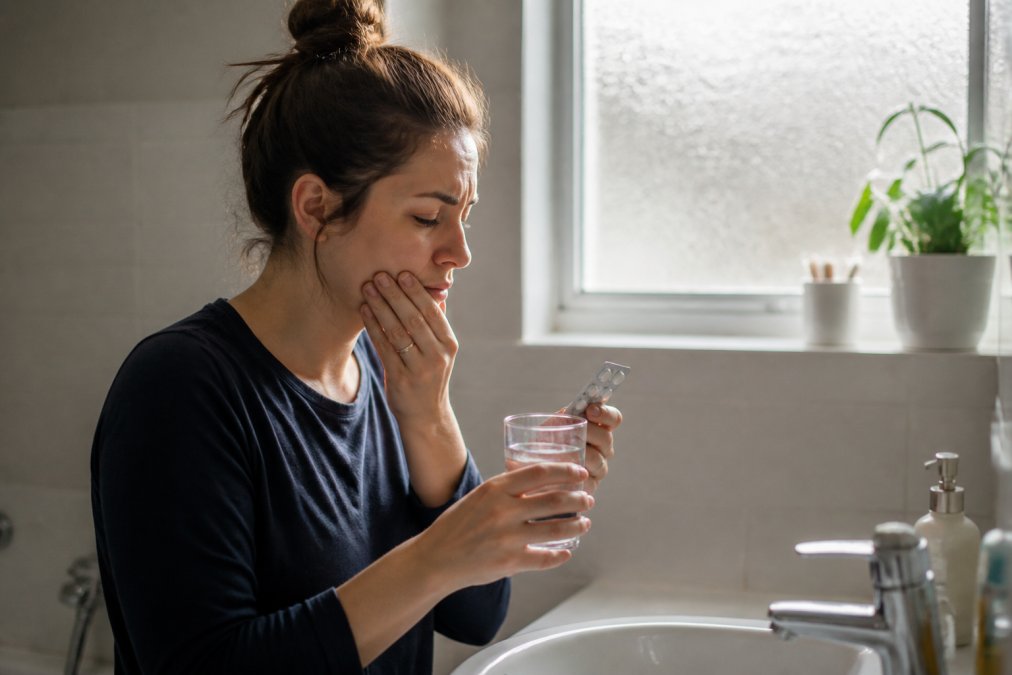 A person at a bathroom sink holding a glass of water and a blister pack of pain relief tablets, soft morning light through a frosted window