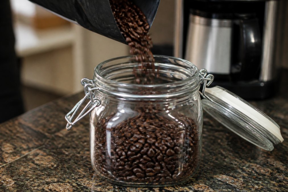 Close-up of fresh coffee beans being poured into a clear glass airtight container with a sealed rubber gasket lid, showing the rich brown color and glossy surface of the beans under natural kitchen lighting