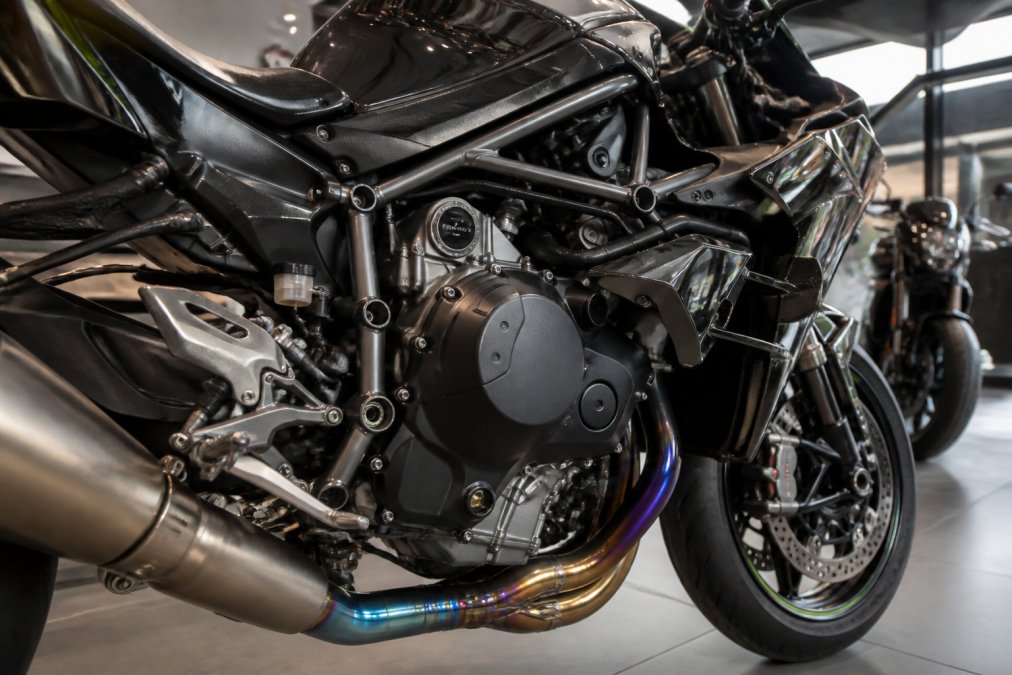Close-up van een Kawasaki H2 superbike in een showroom, met focus op de supercharged motor en het titanium uitlaatsysteem, natuurlijk licht dat de Black Chrome lak accentueert