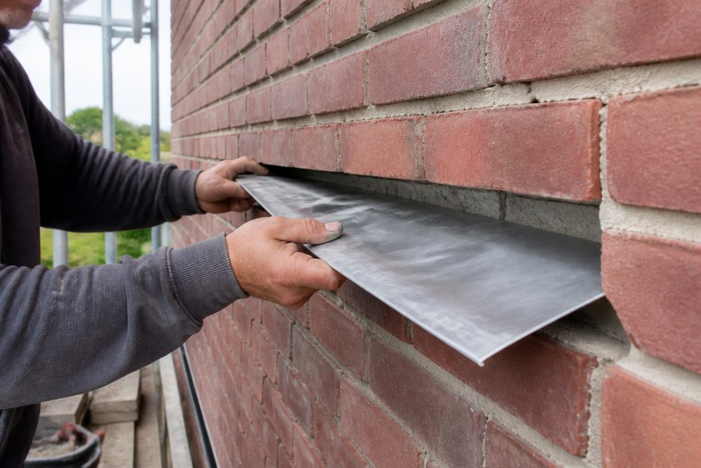 Close-up van een vakman die een strook bladlood in de spouwmuur van een bakstenen gevel plaatst, met zichtbare lintvoeg en metselwerk op de achtergrond