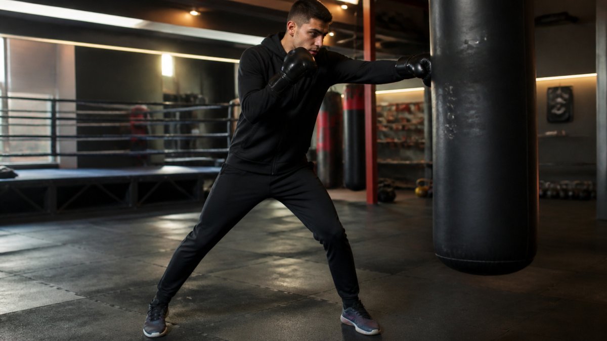 Atleet in zwart Booster trainingspak met capuchon trainingend op punchzak in verlichte boksschool, zichtbaar warm gekleed terwijl hij snelle combinaties uitvoert