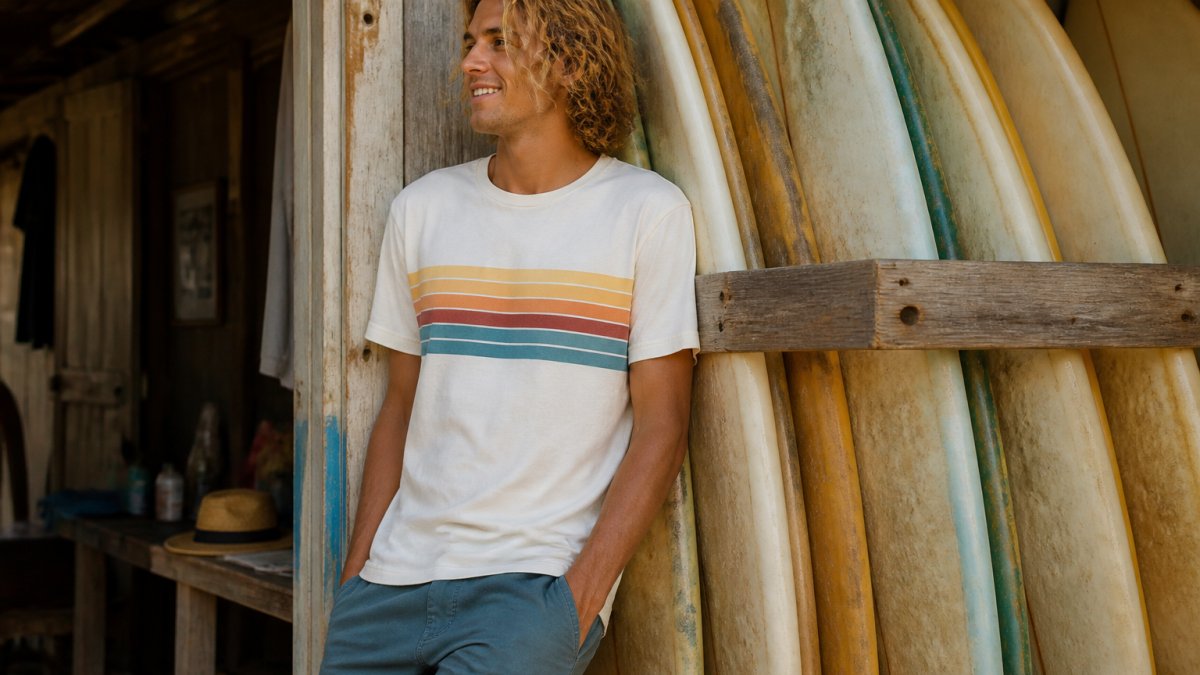 Young adult wearing a retro-striped beach tee leaning against a weathered surfboard rack near a sunny coastal shack