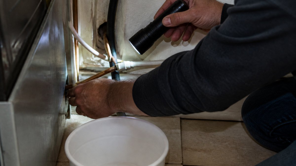 Close-up of a homeowner's hands inspecting the water supply line behind a refrigerator, with a flashlight illuminating the copper or plastic tubing and a bucket nearby