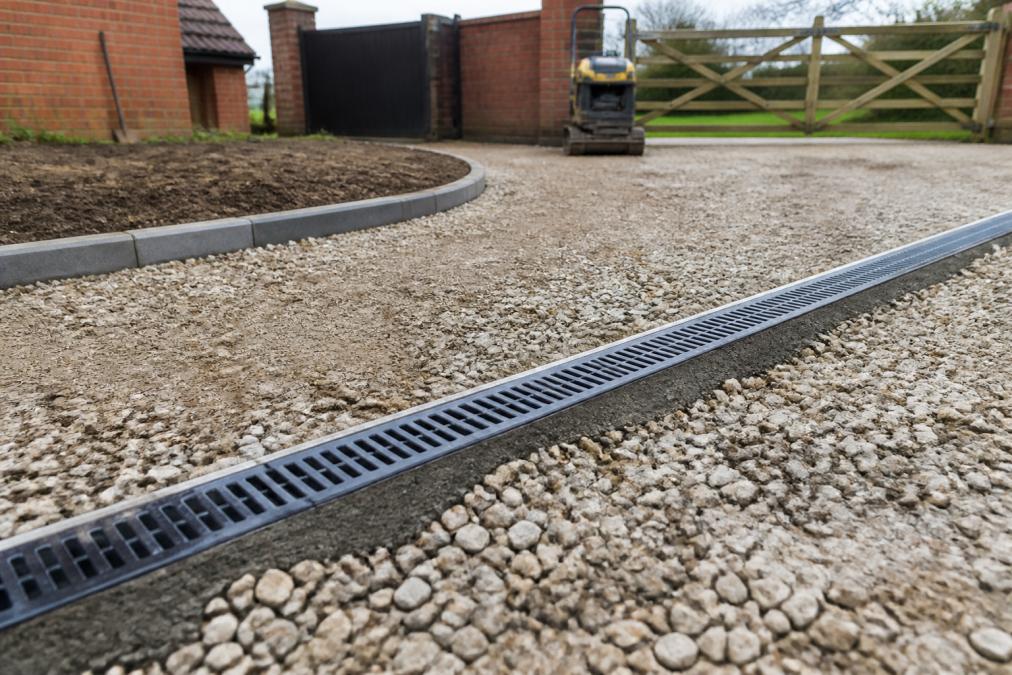 Channel drain system installed across a driveway surface during construction, showing linear drainage grate in place with MOT Type 1 aggregate visible around and beneath it on a Norfolk residential property
