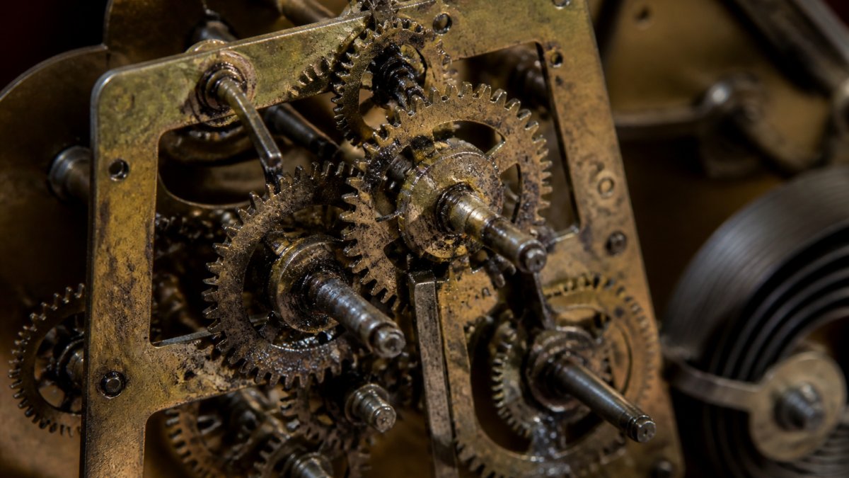 Close-up of a vintage mantle clock's brass gear train with visible dust and dark lubricant residue on the pivots, illuminated by warm workshop light
