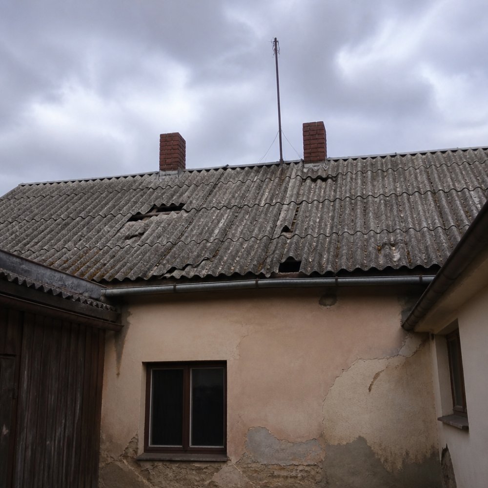 Starší rodinný dům se zvětralou eternitovou střechou, na níž jsou viditelné popraskané a olámané šablony, zatažené nebe, pohled z úrovně dvora