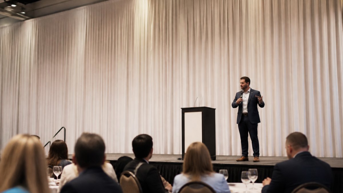 A corporate keynote stage with floor-to-ceiling white pipe and drape backdrop, a clean podium, and a speaker mid-presentation under soft stage lighting