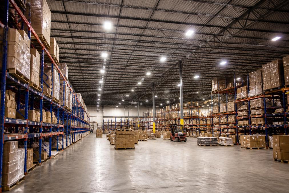 Wide interior shot of a large warehouse with bright LED high bay fixtures illuminating tall metal shelving racks stocked with cardboard boxes, showing clear visibility across the concrete floor under high ceilings