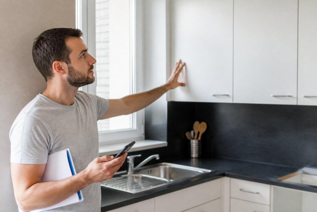 Un acheteur inspectant attentivement les murs, les fenêtres et les installations d'une cuisine pendant une visite immobilière, prenant des notes et des photos avec un smartphone