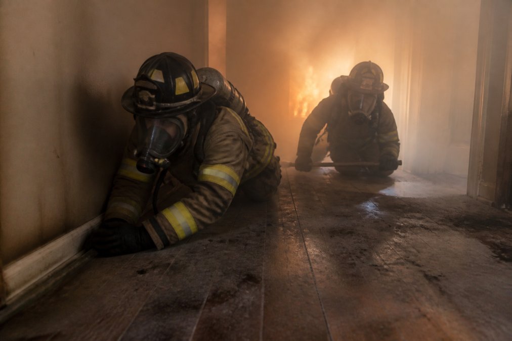 Two firefighters in full turnout gear with SCBA masks crawling low through a smoke-filled training room, one running a gloved hand along the baseboard of a wall while the other sweeps ahead with a tool