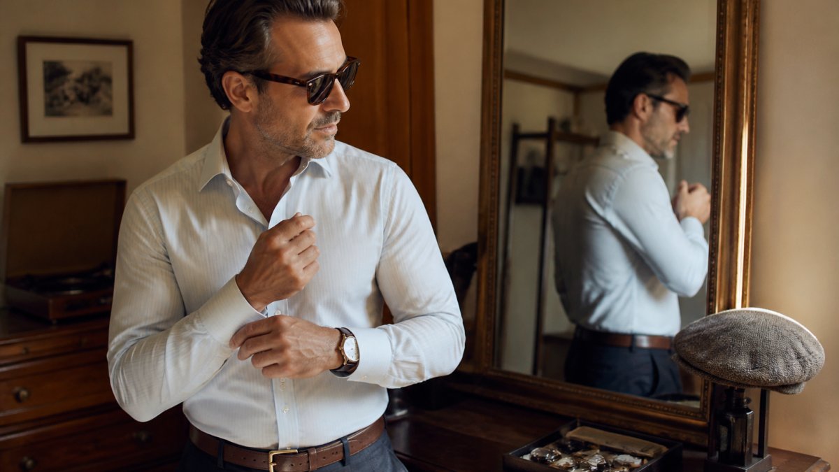 A well-dressed man in his forties wearing a classic watch, leather belt, and retro sunglasses, adjusting his cuff in a warm, softly lit room with a mirror and wooden dresser