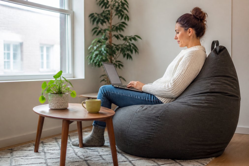 Une personne assise dans un pouf ergonomique de style beanbag dans un coin bureau lumineux, dos bien soutenu, travaillant sur un ordinateur portable posé sur une petite table basse, avec une plante et une tasse de thé à proximité