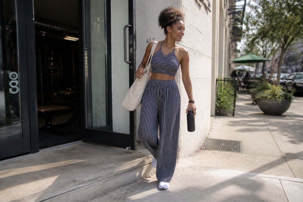 A woman walking from a gym entrance onto a city street wearing a striped halter crop top and wide-leg pants set, carrying a canvas tote bag, morning light, candid stride