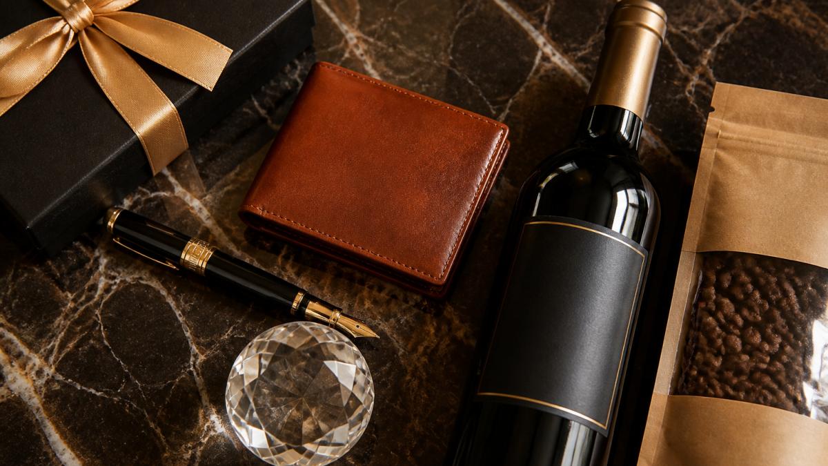 Overhead flat-lay arrangement of luxury corporate gifts including cognac-colored leather wallet, engraved fountain pen, bottle of premium wine, artisanal coffee beans in kraft packaging, and crystal paperweight on dark marble surface with warm golden lighting