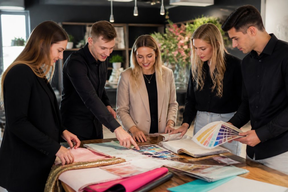 Team van medewerkers in bedrijfsfeest-organisatiebureau die samen decoratiematerialen en huuropties uitzoeken, met catalogi en kleurstalen op tafel onder kantoorverlichting