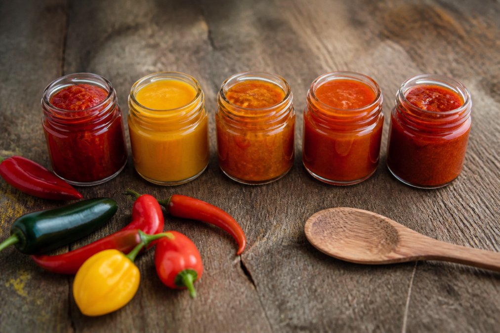 Cinq petits pots de sauces et pâtes de piment artisanales aux couleurs variées (rouge, jaune, orange) disposés sur une table en bois rustique, avec des piments frais et une cuillère en bois à côté
