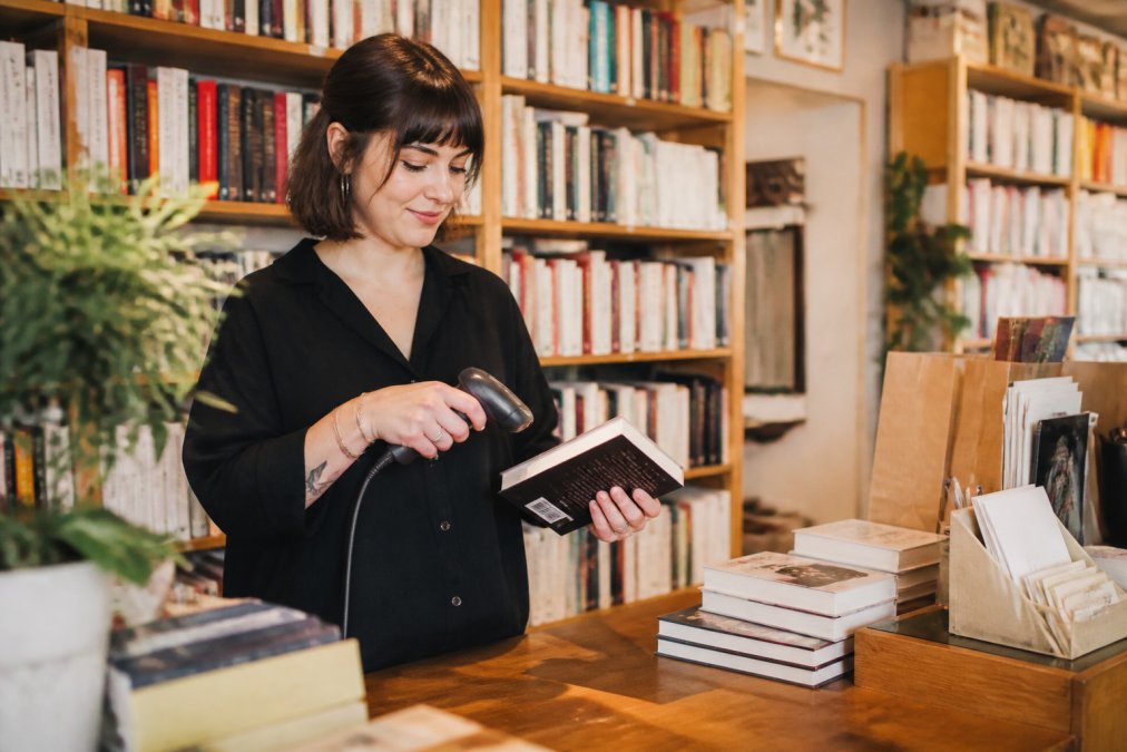 A bookseller at a wooden counter in an independent bookstore scanning a book barcode with a handheld terminal, shelves full of books in the background, warm natural light