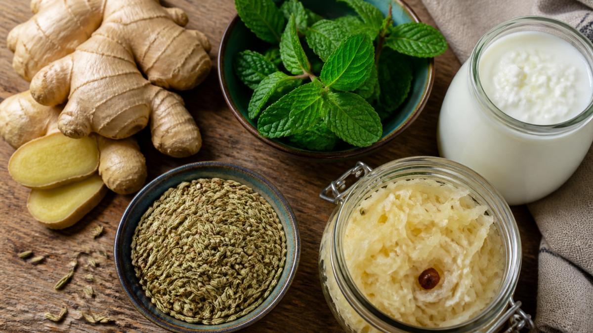 Overhead view of fresh ginger root, peppermint leaves, fennel seeds, and fermented foods like sauerkraut and kefir arranged on a wooden surface with natural lighting